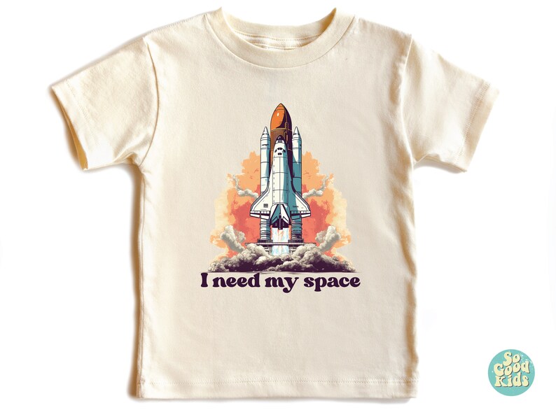 K&ouml;nnte beinhalten: Ein cremefarbenes T-Shirt mit einem Grafikdruck einer Raumf&auml;hre, die in den Himmel startet. Unter dem Grafikdruck steht der Text "I need my space".