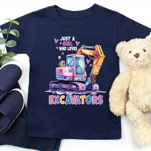 May include: Navy blue toddler t-shirt with a colorful excavator graphic. The design includes the text "JUST A GIRL WHO LOVES EXCAVATORS" with pink hearts. The shirt is paired with navy pants and shoes, and a teddy bear.