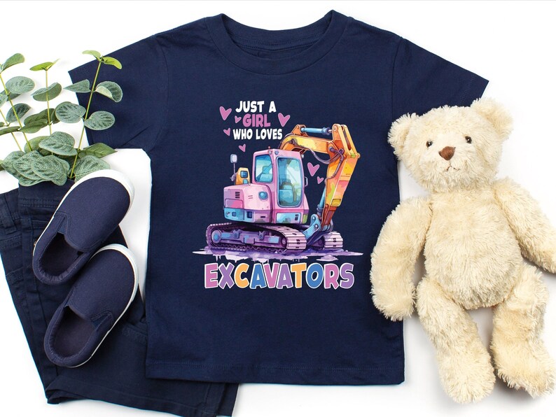 May include: Navy blue toddler t-shirt with a colorful excavator graphic. The design includes the text "JUST A GIRL WHO LOVES EXCAVATORS" with pink hearts. The shirt is paired with navy pants and shoes, and a teddy bear.