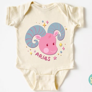 May include: A cream-coloured baby onesie with a pink and blue cartoon ram with the word "Aries" written below it.