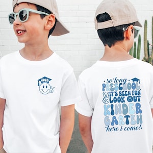 May include: Two white t-shirts with blue text and graphics. The front of one shirt has a smiley face with a graduation cap and the year 2025. The back of the other shirt says "so long preschool it's been fun look out kindergarten here i come!"