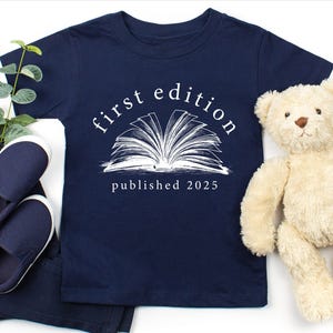 May include: A navy blue t-shirt with a white graphic of an open book and the text "first edition published 2023".