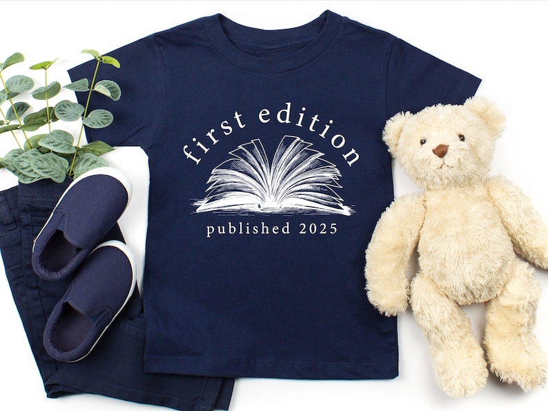 May include: A navy blue t-shirt with a white graphic of an open book and the text "first edition published 2023".