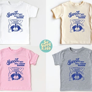 Op de afbeelding: Vier t-shirts in peutermaat met een wasbeer-afbeelding en de tekst "Sweet, but will throw hands". De shirts zijn in wit, beige, roze en grijs.