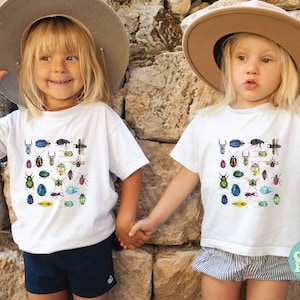 May include: Two children wearing white t-shirts with a colourful insect print design. The shirts feature a variety of beetles, butterflies, and other insects. The children are wearing straw hats and holding hands.