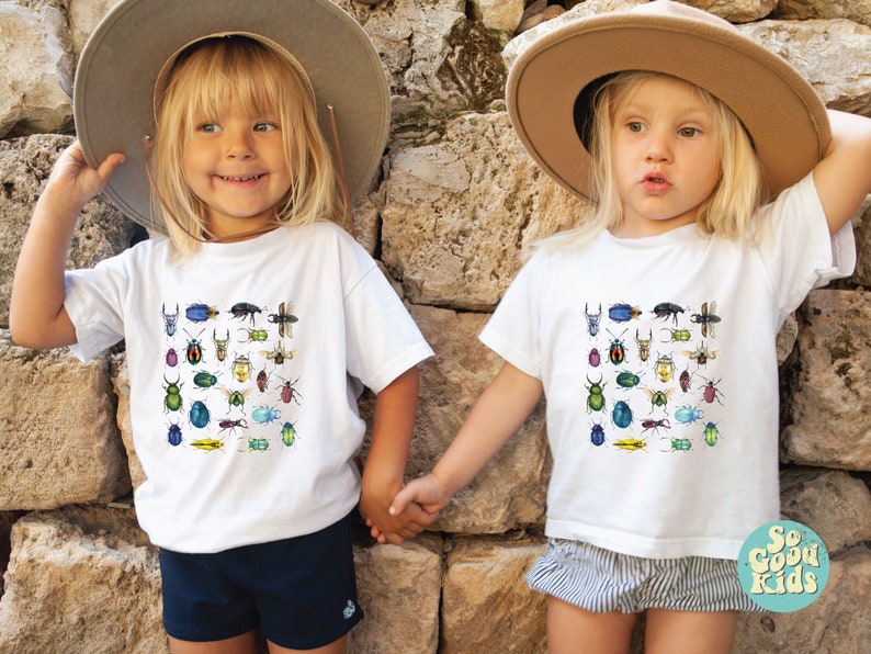 May include: Two children wearing white t-shirts with a colourful insect print design. The shirts feature a variety of beetles, butterflies, and other insects. The children are wearing straw hats and holding hands.