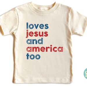 May include: A white t-shirt with the text "loves jesus and america too" in red and blue lettering. The shirt is a toddler size and has a distressed look.