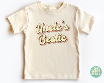 May include: A cream colored short-sleeved t-shirt with the text "Uncle's Bestie" in a retro font with rainbow colored outlines.