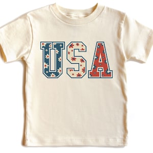 K&ouml;nnte beinhalten: Ein wei&szlig;es T-Shirt mit einer Vintage-Grafik "USA". Die Buchstaben sind mit einem roten, wei&szlig;en und blauen Blumenmuster gef&uuml;llt.