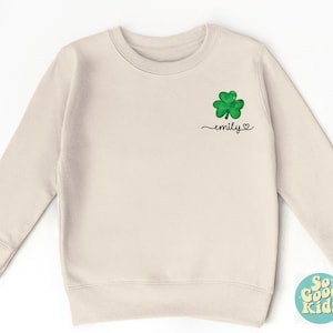 May include: A light beige sweatshirt with a green shamrock and the text "emily" in black.