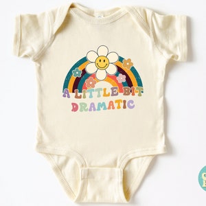 Pode incluir: Um body de beb&ecirc; cor creme com um arco-&iacute;ris e uma flor com um rosto sorridente. O texto "A LITTLE BIT DRAMATIC" &eacute; impresso em letras coloridas.
