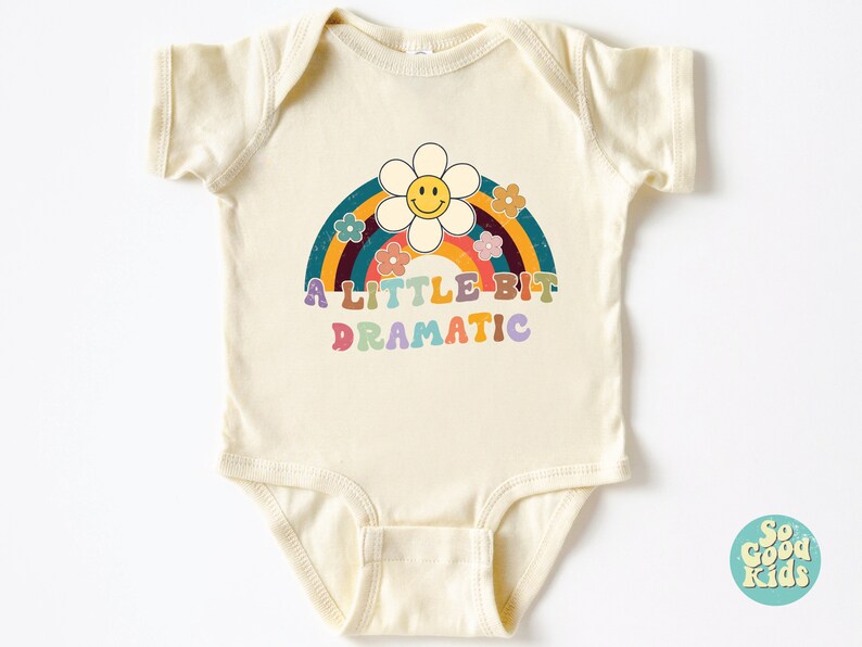 Pode incluir: Um body de beb&ecirc; cor creme com um arco-&iacute;ris e uma flor com um rosto sorridente. O texto "A LITTLE BIT DRAMATIC" &eacute; impresso em letras coloridas.