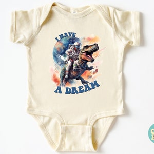 Pode incluir: Um macac&atilde;o de beb&ecirc; de cor creme com um gr&aacute;fico de um astronauta montando um dinossauro no espa&ccedil;o. O texto "I HAVE A DREAM" est&aacute; impresso em letras azuis no macac&atilde;o.