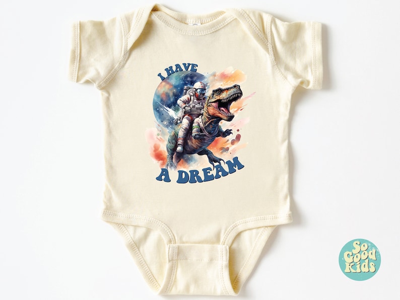 Pode incluir: Um macac&atilde;o de beb&ecirc; de cor creme com um gr&aacute;fico de um astronauta montando um dinossauro no espa&ccedil;o. O texto "I HAVE A DREAM" est&aacute; impresso em letras azuis no macac&atilde;o.