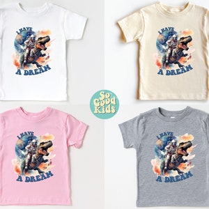 Pode incluir: Quatro camisetas para crian&ccedil;as pequenas em branco, bege, rosa e cinza com uma estampa gr&aacute;fica de um astronauta montando um dinossauro. O gr&aacute;fico tem um efeito de aquarela e o texto "I HAVE A DREAM" &eacute; impresso abaixo da imagem.