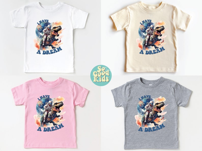 Pode incluir: Quatro camisetas para crian&ccedil;as pequenas em branco, bege, rosa e cinza com uma estampa gr&aacute;fica de um astronauta montando um dinossauro. O gr&aacute;fico tem um efeito de aquarela e o texto "I HAVE A DREAM" &eacute; impresso abaixo da imagem.