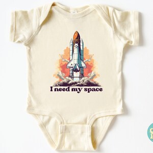 K&ouml;nnte beinhalten: Ein cremefarbener Baby-Strampler mit einer Grafik eines Raumschiffs, das in den Himmel startet. Der Text "I need my space" ist unter dem Bild gedruckt.