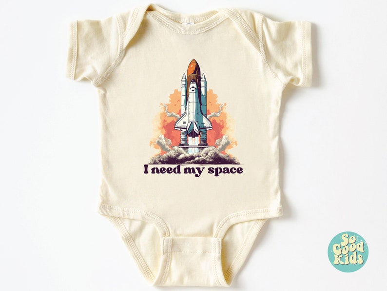 K&ouml;nnte beinhalten: Ein cremefarbener Baby-Strampler mit einer Grafik eines Raumschiffs, das in den Himmel startet. Der Text "I need my space" ist unter dem Bild gedruckt.