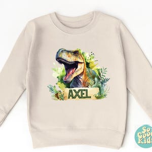 May include: A beige sweatshirt featuring a watercolor illustration of a Tyrannosaurus Rex with an open mouth, surrounded by green foliage. The name "AXEL" is printed on a banner below the dinosaur. The sweatshirt is designed for kids.