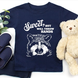 Op de afbeelding: Een marineblauw t-shirt met een witte afbeelding van een wasbeer met de tekst "Sweet, but will throw hands".
