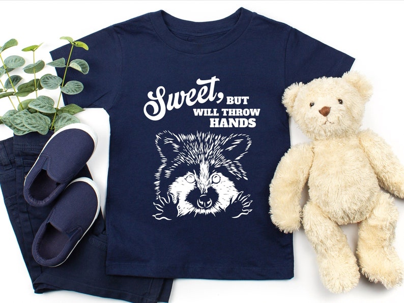 Op de afbeelding: Een marineblauw t-shirt met een witte afbeelding van een wasbeer met de tekst "Sweet, but will throw hands".