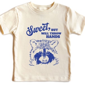 Op de afbeelding: Een cr&egrave;mekleurig t-shirt met een blauwe wasbeer afbeelding en de tekst "Sweet, but will throw hands".