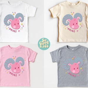 May include: Four toddler-sized t-shirts in white, beige, pink, and grey with a pink cartoon ram head with blue horns and the word "Aries" printed on each shirt.