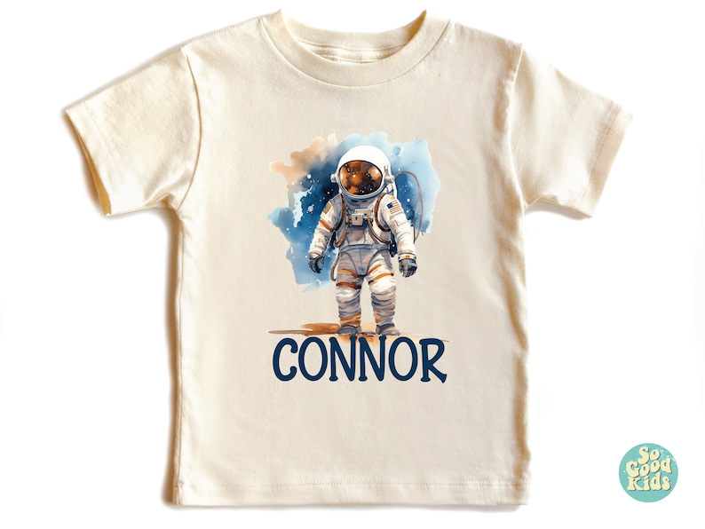 May include: A cream-colored t-shirt with a watercolor illustration of an astronaut in a space suit standing on a planet. The text "CONNOR" is printed below the astronaut.