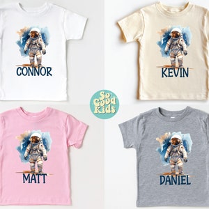 May include: Four personalized t-shirts featuring a watercolor illustration of an astronaut on a planet with the names Connor, Kevin, Matt, and Daniel printed below.