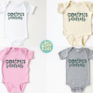 May include: Four baby onesies in different colors: white, cream, pink, and gray. Each onesie has the text "COUSINsaur" printed in dark green with a dinosaur silhouette graphic.