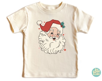 May include: A cream colored t-shirt with a vintage illustration of Santa Claus's face. The illustration is in red, white, and green with pink sparkles around the image. The text "So Good Kids" is in the bottom right corner of the image.