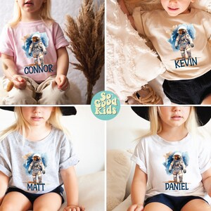 May include: Four children's t-shirts with an astronaut graphic and personalized names. The shirts are in pink, beige, grey, and white. The names on the shirts are Connor, Kevin, Matt, and Daniel.