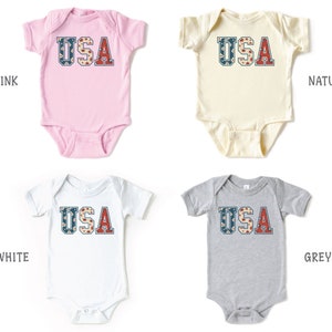 K&ouml;nnte beinhalten: Vier Baby-Bodys in verschiedenen Farben mit dem Wort "USA" auf der Vorderseite. Die Bodys sind in Pink, Wei&szlig;, Natur und Grau.