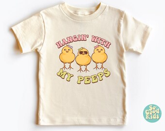 Peut inclure: Un t-shirt crème avec un motif de trois poussins jaunes portant des nœuds et des lunettes de soleil. Le texte "Hangin' with my peeps" est imprimé en lettres roses et vertes.