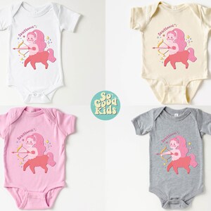 May include: Four baby onesies in different colours, white, beige, pink, and grey, with a pink cartoon Sagittarius zodiac sign with an arrow and the text "Sagittarius" on each onesie.