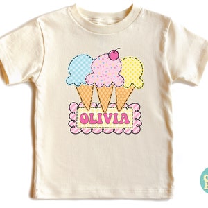 May include: A light cream colored t-shirt with a graphic of three ice cream cones with a cherry on top. The ice cream cones are in blue, pink, and yellow. The text "OLIVIA" is on a pink and white striped banner.