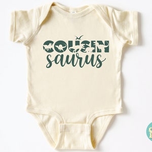 May include: A cream-colored baby onesie with a green graphic that says "COUSIN saurus" with dinosaur silhouettes.