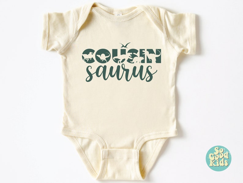 May include: A cream-colored baby onesie with a green graphic that says "COUSIN saurus" with dinosaur silhouettes.