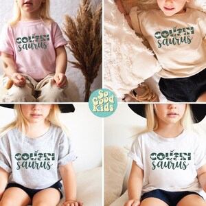 May include: Four children wearing t-shirts with the text "Cousin-saurus" printed on them. The shirts are in different colors: pink, beige, gray, and white. The children are wearing shorts and are sitting on a white surface.