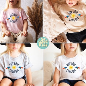 May include: A collection of children's t-shirts in various colors, each featuring the phrase "SUN'S OUT GUNS OUT" with a sun and water gun graphic. The shirts are light pink, beige, gray, and white.