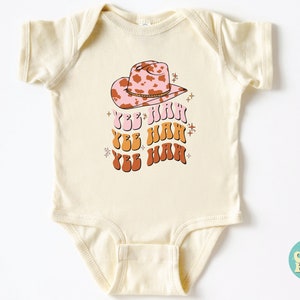 K&ouml;nnte beinhalten: Ein cremefarbener Baby-Strampler mit einem rosa und braunen Cowboyhut mit Kuhmuster und dem Text "Yee Haw", der dreimal in einer Retro-Schrift wiederholt wird.