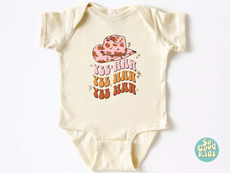 K&ouml;nnte beinhalten: Ein cremefarbener Baby-Strampler mit einem rosa und braunen Cowboyhut mit Kuhmuster und dem Text "Yee Haw", der dreimal in einer Retro-Schrift wiederholt wird.