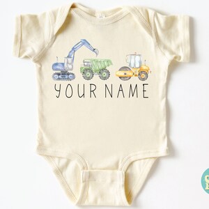 May include: A cream-colored baby onesie with a graphic of three construction vehicles: a blue excavator, a green dump truck, and a yellow road roller. The text "YOUR NAME" is printed below the vehicles.