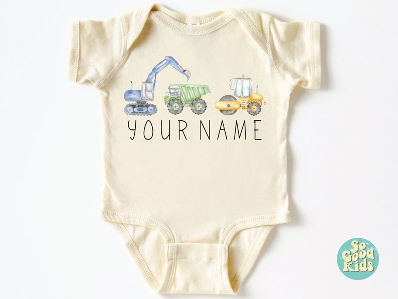 May include: A cream-colored baby onesie with a graphic of three construction vehicles: a blue excavator, a green dump truck, and a yellow road roller. The text "YOUR NAME" is printed below the vehicles.