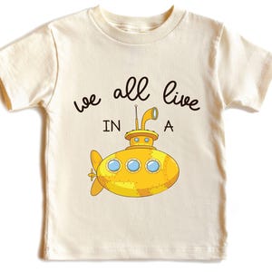 May include: Cream-colored t-shirt featuring a yellow submarine illustration with the text "we all live in a". The submarine has portholes and a periscope. The shirt is designed for kids.