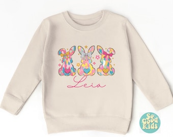May include: A cream-colored children's sweatshirt featuring three colorful bunny designs with bows and hearts. The name "Leia" is written in pink script below the bunnies. The sweatshirt has long sleeves and a round neckline.