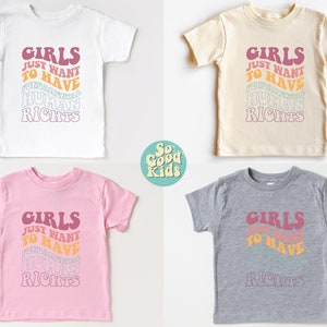May include: Four toddler-sized t-shirts in white, beige, pink, and gray with the text "Girls Just Want To Have Fundamental Human Rights" in a retro font with a rainbow color scheme.