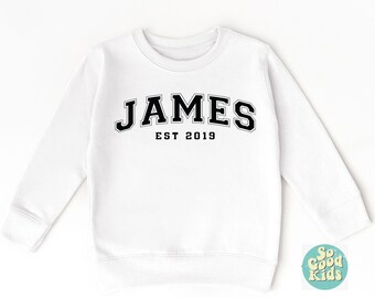 May include: White sweatshirt with the name "JAMES" in black varsity-style lettering, with "EST 2019" below. The sweatshirt has a crew neck and long sleeves. A small logo is visible in the bottom right corner.