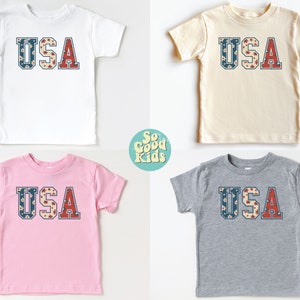 K&ouml;nnte beinhalten: Vier T-Shirts in Kindergr&ouml;&szlig;e mit dem Wort "USA" auf der Vorderseite. Die Shirts sind in Wei&szlig;, Beige, Rosa und Grau. Die Buchstaben bestehen aus einem roten, wei&szlig;en und blauen Blumenmuster.