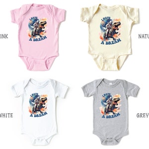 Pode incluir: Quatro bodies de beb&ecirc; em diferentes cores: rosa, natural, branco e cinza. Cada body apresenta um gr&aacute;fico de um astronauta montando um dinossauro com o texto "Eu tenho um sonho".